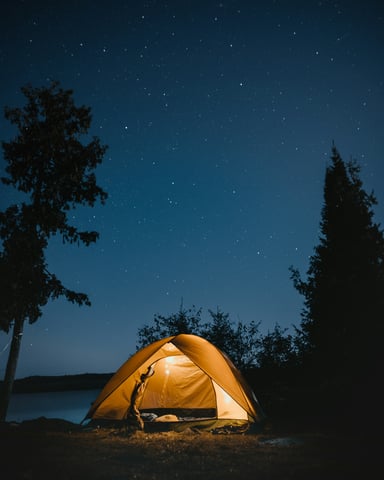 Illuminated yellow tent on lakeside campground surrounded by evergreen trees under starry night sky