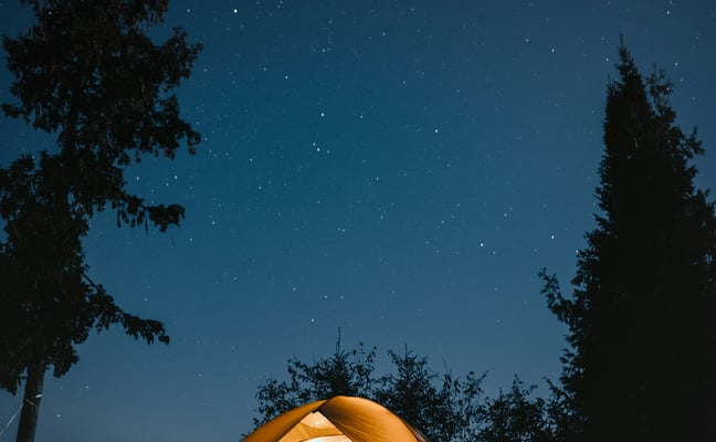 Illuminated yellow tent on lakeside campground at night under starry sky with evergreen trees