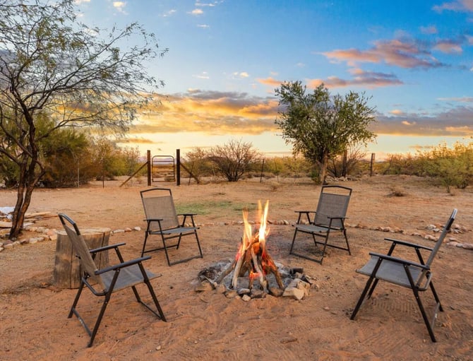 Desert campsite with burning campfire, four camping chairs, and gate at sunset